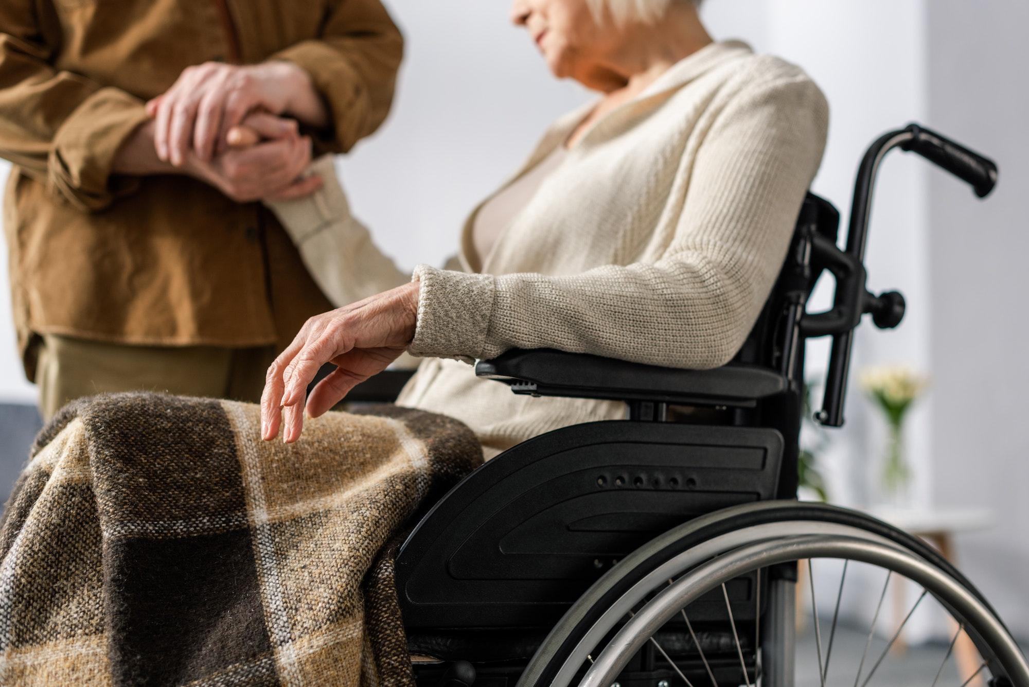 cropped view of senior man holding hand of handicapped, ill wife in wheelchair